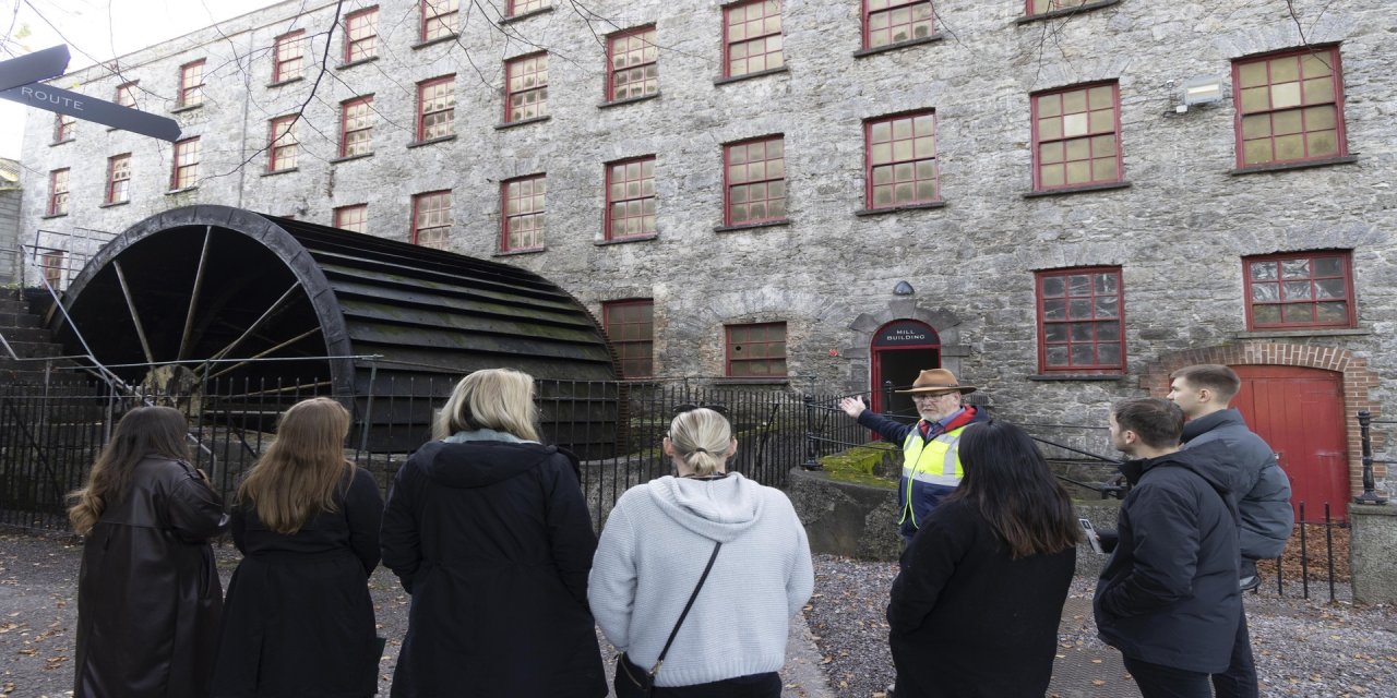 Five women and two men standing outside the grey stoned building of Midleton Distillery. Tour guide wearing a hat is explaining the history while pointing at the building. Large, black barrell to the left of the red outlined windows. 