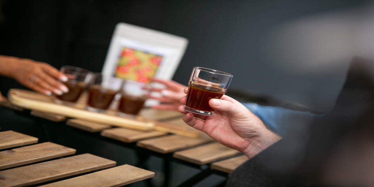 Brown liquid in a short glass in a mans hand. Wooden table and white picture frame in the background 