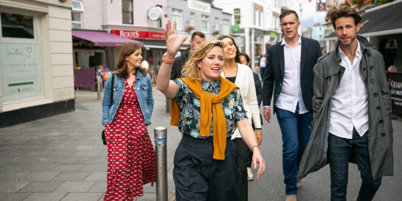 Group of people following tour guide wearing yellow jumper around her neck with blonde curly hair